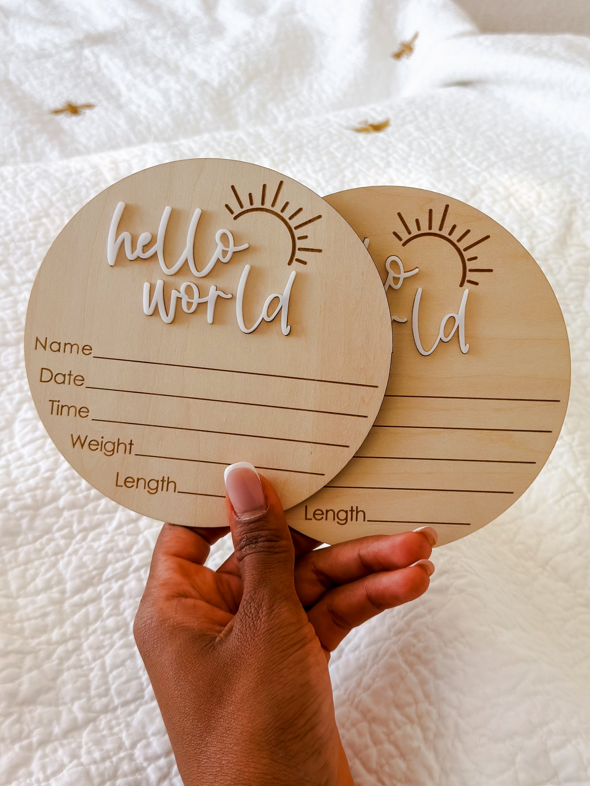 Two wooden circular baby milestone cards with 'hello world' text held by a hand on a white background.