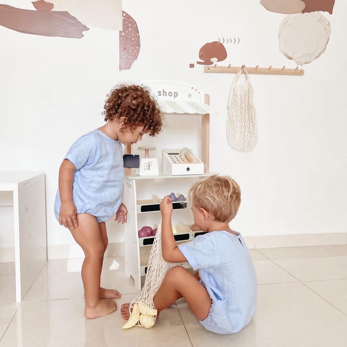 Two toddlers in matching blue rompers playing with a wooden play shop, filling a cotton net shopping bag with wooden fruit.