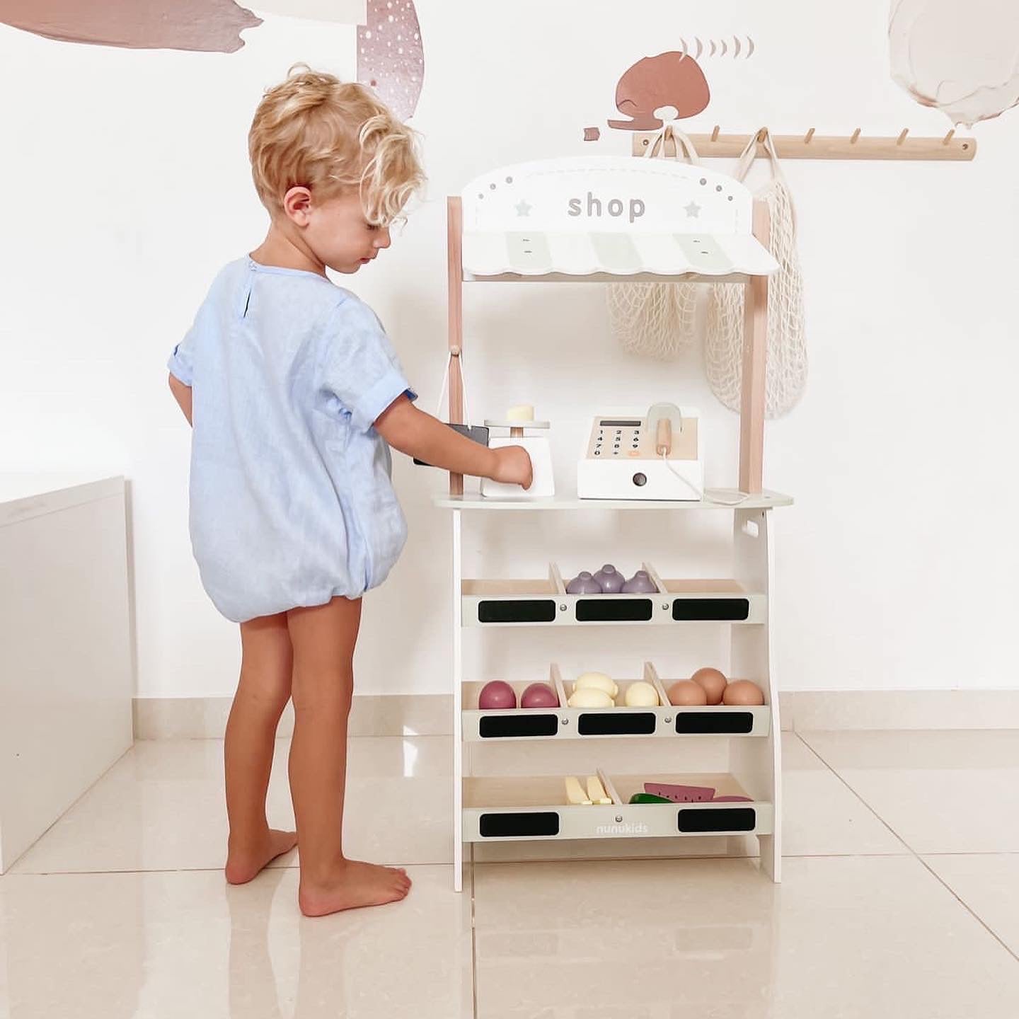 Young child in blue romper pretending to shop with a Maxim’s wooden play shop, exploring the wooden cash register and play food.
