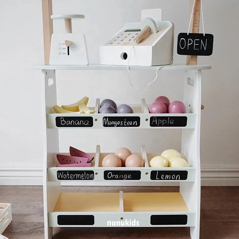 Wooden play shop with sage green awning, cash register, wooden fruit and chalkboard labels, displayed in a modern Scandi-style nursery.